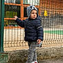 child, kid, hat, jacket, fence, outdoor, shoes, sneakers, jeans, playground, building, window, side_view, standing, cold_weather, casual_clothing, curious, young, person, urban