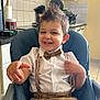child, toddler, smiling, high_chair, bow_tie, suspenders, white_shirt, brown_shorts, kitchen, countertop, paper_towel, utensils, tile_wall, cabinet, happy, indoor, person, cute, playful, hair_spikes