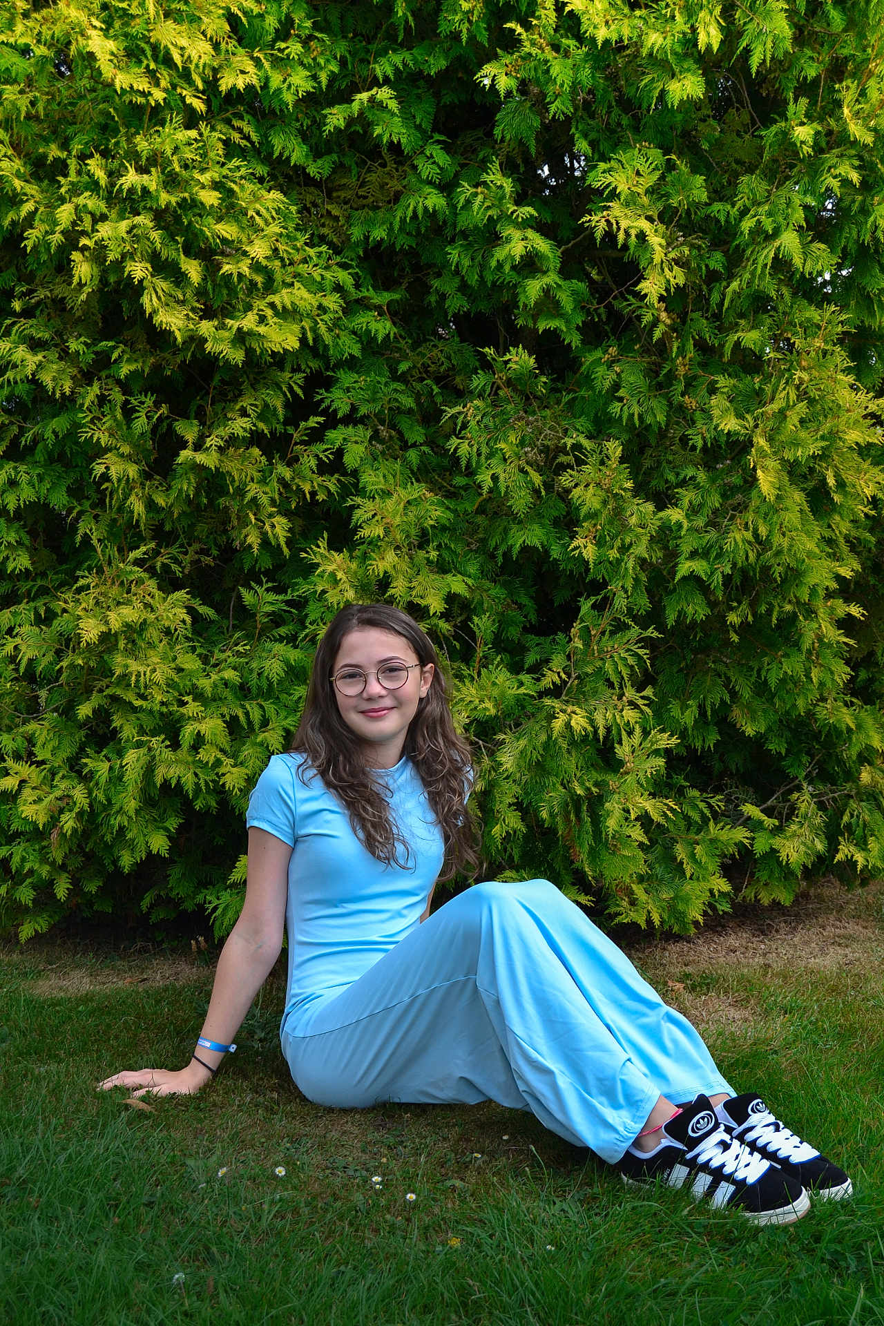 Sarah a rejoint le concours — aidez-le/la à gagner de superbes lots ! girl, child, glasses, blue_clothing, grass, greenery, bush, outdoor, casual, sitting, smile, shoes, sneakers, young, nature, portrait, daylight, relaxed, happy, fashion