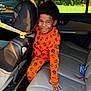 child, smiling, orange_pajamas, pumpkin_pattern, car_interior, backseat, seatbelt, headrest, sunlight, happy, young_boy, window, seat, cookies, oreo, leather_seat, playful, casual, indoors, portrait
