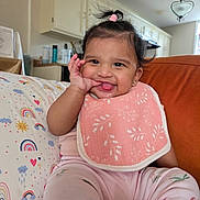 Amira joined the competition — help win amazing prizes! baby, child, infant, smiling, happy, tongue_out, portrait, sitting, sofa, couch, cushion, pink_clothing, bib, hair_bow, ponytail, earring, indoor, kitchen, home, rainbow_pattern