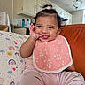 baby, child, infant, smiling, happy, tongue_out, portrait, sitting, sofa, couch, cushion, pink_clothing, bib, hair_bow, ponytail, earring, indoor, kitchen, home, rainbow_pattern