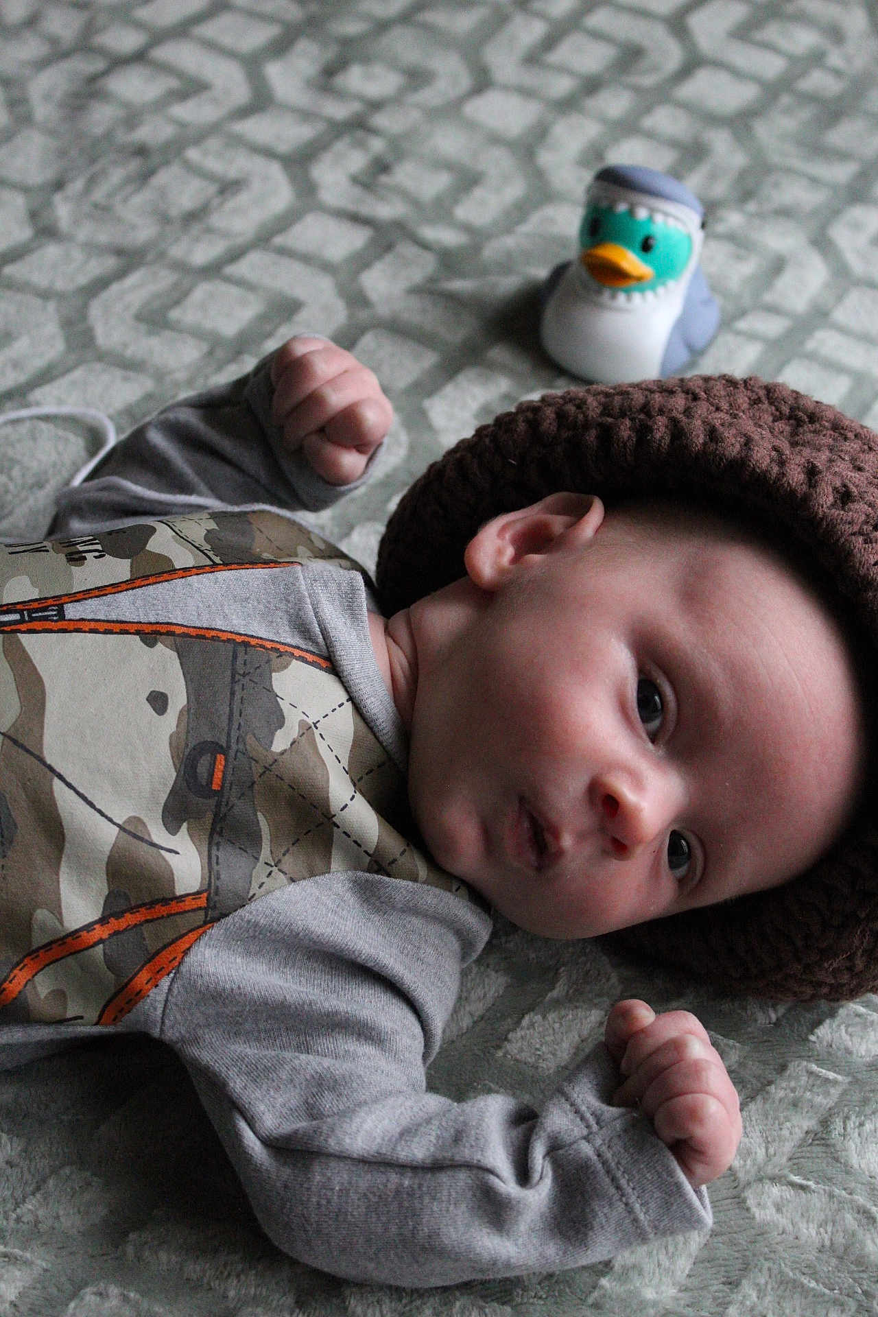 Oakley joined the competition — help win amazing prizes! baby, infant, child, hat, knitted_hat, camouflage_clothing, toy_duck, blanket, texture, fist, curious_expression, lying_down, cute, indoor, soft_lighting, face, head, person, small_hands, playful