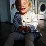 Eva a rejoint le concours — aidez-le/la à gagner de superbes lots ! toddler, child, smiling, pigtails, sweater, cat_design, indoor, floor, kitchen, refrigerator, washing_machine, sunlight, shadow, happy, cute, seated, legs, clothing, home, playful