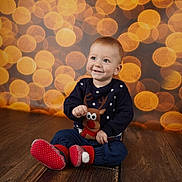 Kaloyan is registered to the contest to win money with this photo: baby, background_lights, bokeh, child, cozy, cute, festive, happy, holiday, indoor, infant, playful, portrait, red_slippers, reindeer_sweater, sitting, smiling, warm_lighting, winter_clothing, wooden_floor