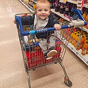 Kaloyan is registered to the contest to win money with this photo: aisle, baby, bottle, child, clothing, drink, floor, footwear, grocery, handle, happy, packaging, person, retail, shelf, shopping_cart, sitting, smile, store, supermarket
