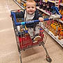 Kaloyan is registered to the contest to win money with this photo: aisle, baby, bottle, child, clothing, drink, floor, footwear, grocery, handle, happy, packaging, person, retail, shelf, shopping_cart, sitting, smile, store, supermarket
