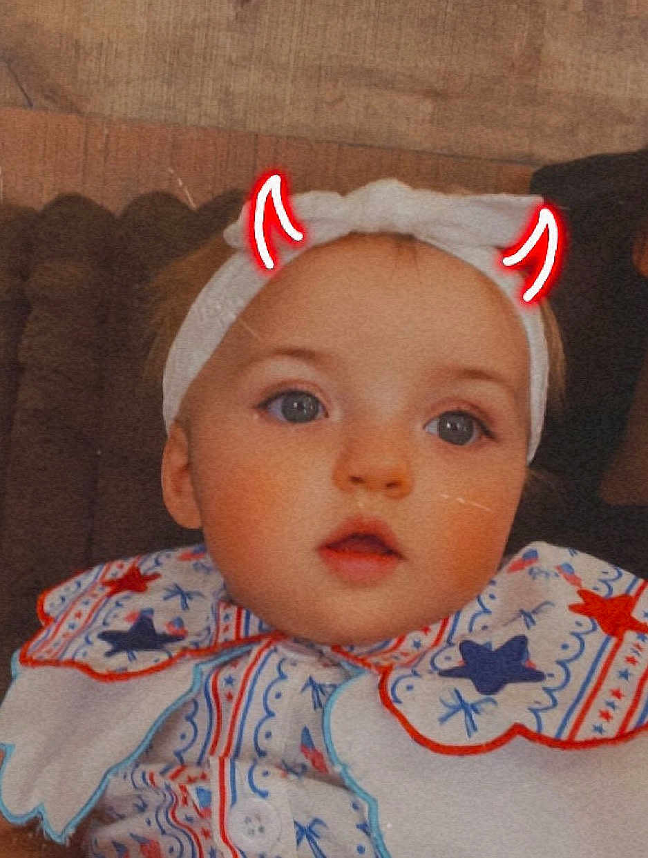 Ashlyn Rae joined the competition — help win amazing prizes! baby, bib, blue_eyes, child, close_up, couch, cute, doodle, expression, face, headband, headwear, indoors, infant, patterned_clothing, portrait, red_horns, seated, soft_lighting, young_child