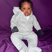 Mason is registered to the contest to win money with this photo: baby, infant, child, onesie, gray_clothing, mittens, purple_background, sitting, drool, curly_hair, portrait, indoor, cozy, casual, cute, young_child, face, expression, person, apparel
