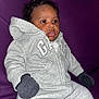 baby, child, hoodie, mittens, gray_clothing, sitting, purple_background, curly_hair, indoor, portrait, casual, cute, young_child, warm_clothing, face, expression, cozy, infant, person, comfort