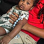 baby, child, infant, dinosaur_onesie, pacifier_clip, blanket, couch, holding, curly_hair, red_shirt, beige_pants, indoors, close_up, comfortable, cute, relaxed, skin, hand, face, portrait