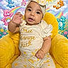 baby, child, infant, yellow_chair, headband, smile, waving_hand, care_bears, rainbow, sun, clouds, flowers, cartoon_background, plush_chair, cute, outfit, patterned_pants, happy, portrait, indoor
