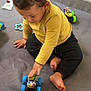 toddler, child, toy_car, blue_wheels, yellow_shirt, black_pants, play_mat, indoor, playing, curly_hair, barefoot, hand, sitting, focus, colorful, plastic_toy, baby, floor, playpen, crawling_area