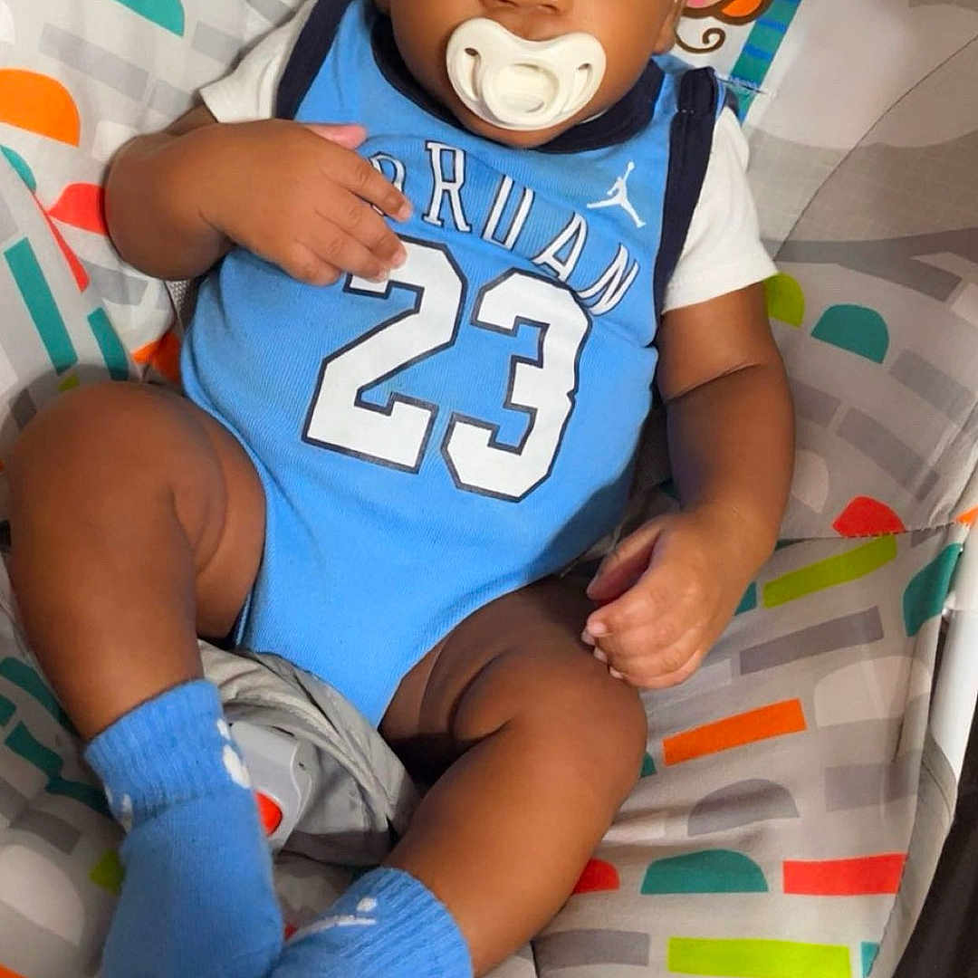 Zareese Stouton is registered to the contest to win money with this photo: baby, child, infant, portrait, sitting, lying_down, pacifier, smiling, jersey, basketball_jersey, number_23, blue_clothing, socks, colorful, playmat, patterned_background, indoor, baby_seat, cozy, face