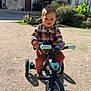 child, toddler, tricycle, outdoor, sunny, plaid_shirt, brown_pants, shoes, playground, bench, building, window, greenery, sidewalk, toy, person, happy, daylight, street, urban