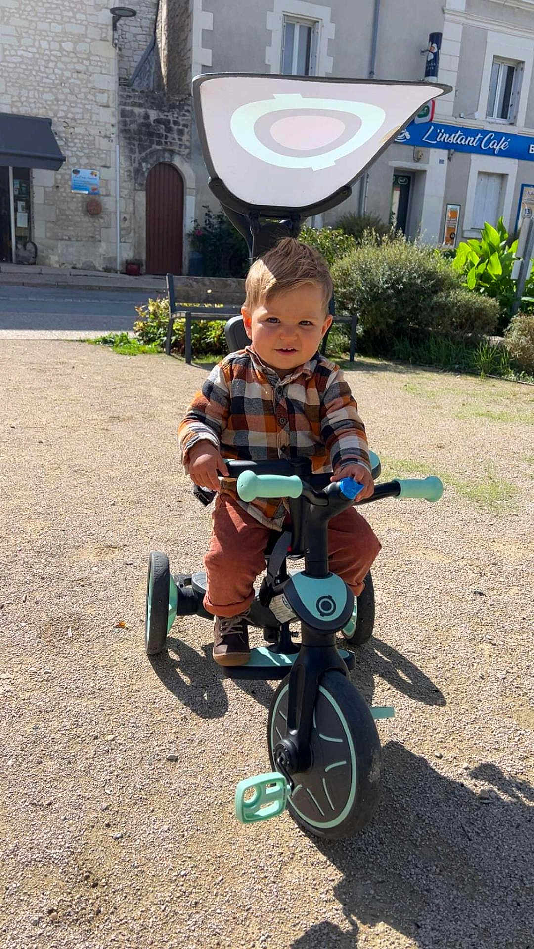 Evan participe au concours pour gagner de l'argent avec cette photo : child, toddler, tricycle, outdoor, sunny, plaid_shirt, brown_pants, shoes, playground, bench, building, window, greenery, sidewalk, toy, person, happy, daylight, street, urban