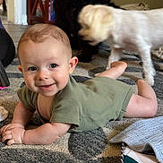 Leon is registered to the contest to win money with this photo: baby, child, infant, green_onesie, carpet, floor, dog, pet, blurred_motion, indoor, smiling, face, hands, legs, home, furniture, blanket, colorful_bag, playful, crawling