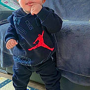 Thyam participe au concours pour gagner de l'argent avec cette photo : toddler, child, standing, indoors, clothing, hoodie, sneakers, jordan, black_clothing, red_accent, floor_mat, couch, blanket, curious, finger_in_mouth, young_child, casual_wear, baby, footwear, person