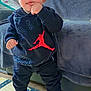 toddler, child, standing, indoors, clothing, hoodie, sneakers, jordan, black_clothing, red_accent, floor_mat, couch, blanket, curious, finger_in_mouth, young_child, casual_wear, baby, footwear, person