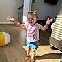 toddler, child, barefoot, curly_hair, pink_shirt, blue_shorts, indoor, wooden_floor, sunlight, beach_ball, smiling, arms_outstretched, home, living_room, mirror, television, rug, basket, furniture, playful