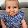 baby, child, blue_eyes, smile, dress, floral_pattern, couch, indoor, cute, happy, portrait, infant, sitting, soft_lighting, human, person, short_hair, plump_cheeks, cozy, casual