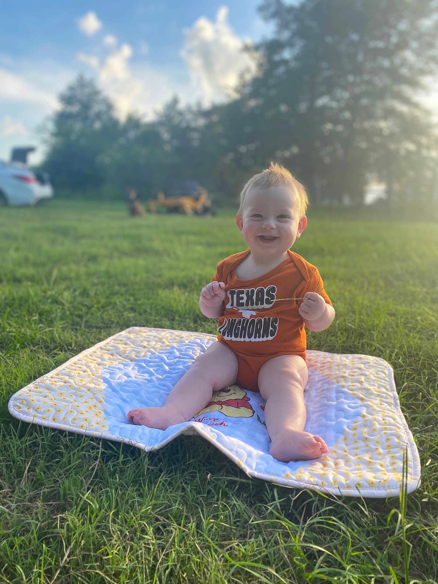 Jasper is registered to the contest to win money with this photo: baby, child, grass, outdoor, sunlight, quilt, onesie, smiling, happy, grass_field, nature, daylight, person, infant, clothing, greenery, cute, sitting, playful, summer