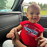 Daniel is registered to the contest to win money with this photo: baby, child, smiling, red_shirt, car, car_interior, window, rain, hand, adult, holding, seat, greenery, daylight, portrait, cute, happy, person, clothing, indoors