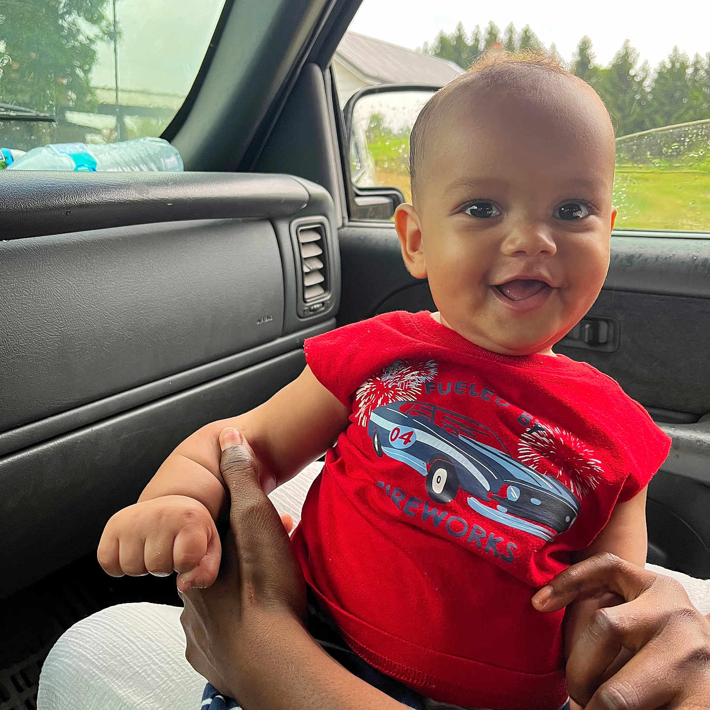 Daniel is registered to the contest to win money with this photo: baby, child, smiling, red_shirt, car, car_interior, window, rain, hand, adult, holding, seat, greenery, daylight, portrait, cute, happy, person, clothing, indoors
