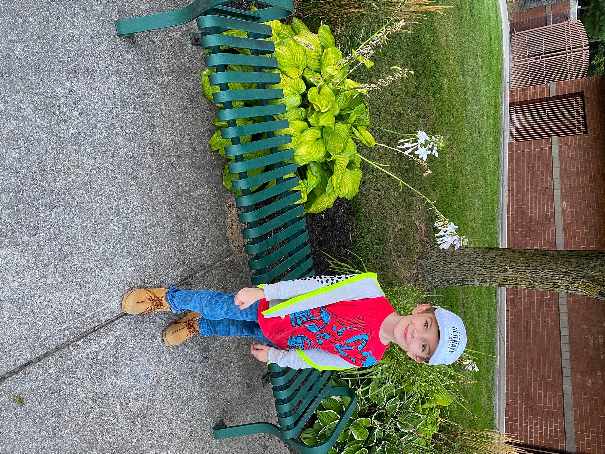 Mark is registered to the contest to win money with this photo: asphalt, concrete, flower, flowering_plant, grass, green, hat, headwear, herb, joy, line, person, plant, plant_stem, shadow, shrub, sidewalk, smile, terrestrial_plant, toddler