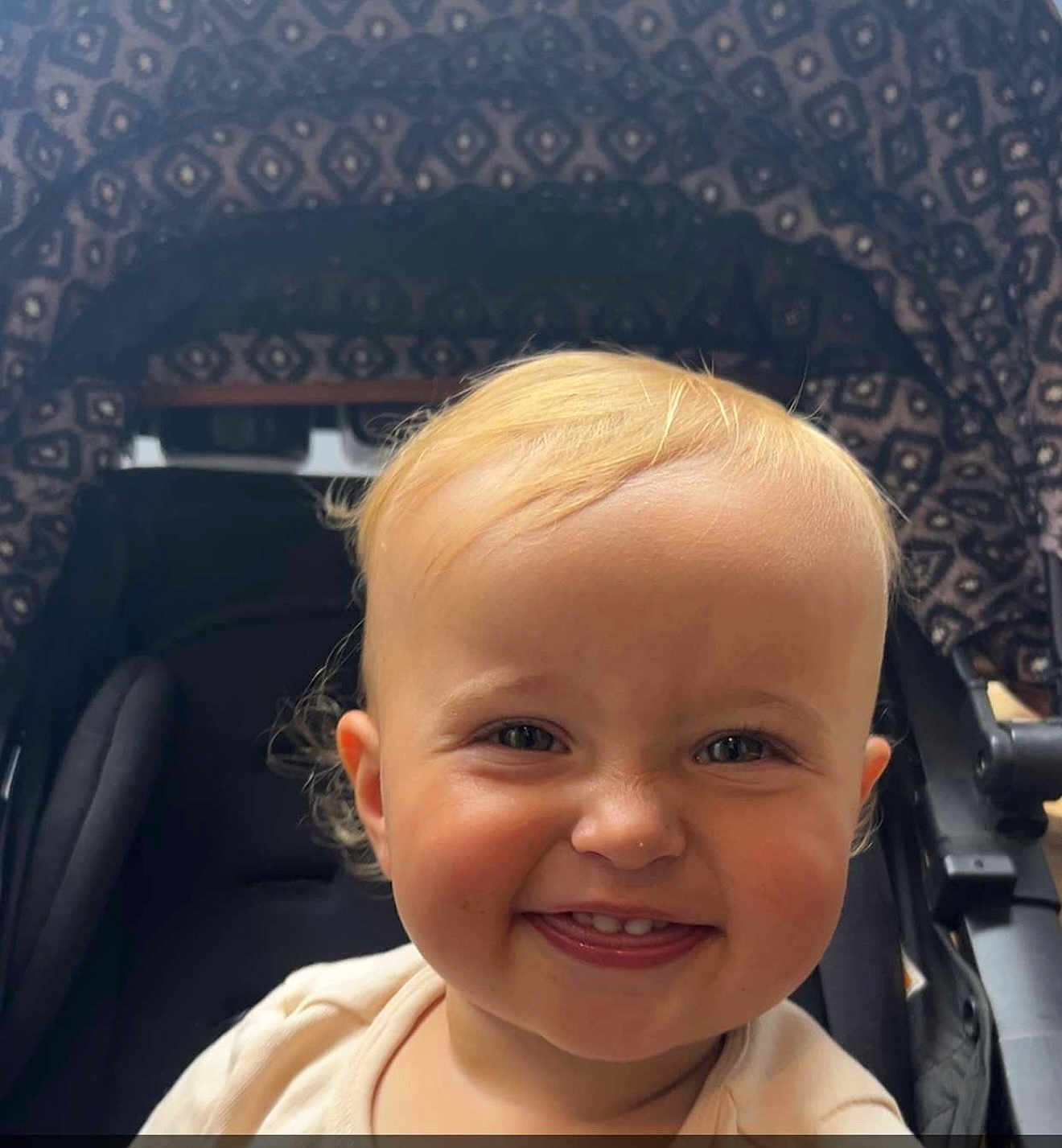 Payton is registered to the contest to win money with this photo: baby, child, toddler, smiling, face, blonde_hair, teeth, stroller, seat, pattern, canopy, happy, cute, infant, person, portrait, closeup, outdoor, daylight, clothing