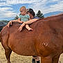 Payton is registered to the contest to win money with this photo: child, toddler, horse, animal, outdoor, grass, person, adult, sky, clouds, mountains, nature, footwear, hat, hand, barefoot, brown_horse, casual_clothing, daytime, support
