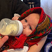 Iris is registered to the contest to win money with this photo: baby, babywearing, bottle, closeup, clothing, cute, eyes, feeding, feeding_time, hand, headscarf, indoors, infant, milk, patterned_cloth, person, portrait, red, soft_light, traditional