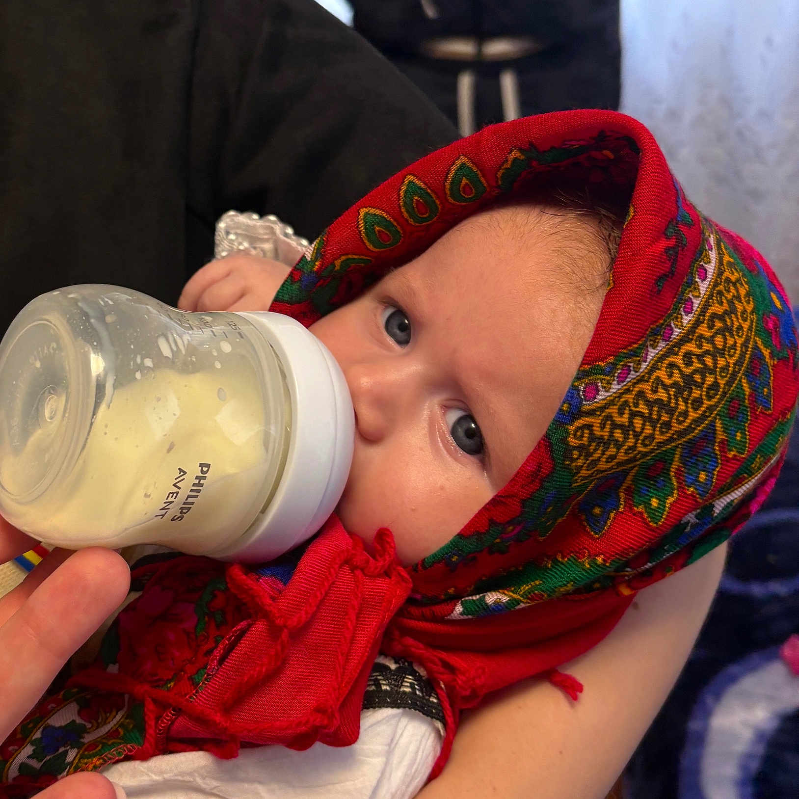 Iris is registered to the contest to win money with this photo: baby, babywearing, bottle, closeup, clothing, cute, eyes, feeding, feeding_time, hand, headscarf, indoors, infant, milk, patterned_cloth, person, portrait, red, soft_light, traditional