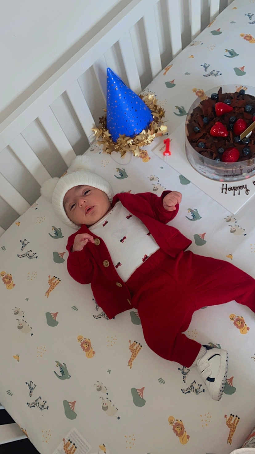 Rahim is registered to the contest to win money with this photo: baby, crib, birthday_cake, party_hat, red_clothing, white_hat, animal_print, giraffe, zebra, lion, crocodile, strawberries, blueberries, cake, celebration, first_birthday, infant, footwear, bed, sleepwear