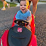 baby, bib, blue_shirt, child, driveway, face, family, happy, outdoor, parked_car, playtime, portrait, red_car, smiling, steering_wheel, summer, sunlight, toddler, toy_car, vehicle