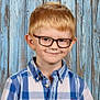 background, blue_shirt, boy, buttoned_shirt, casual_clothing, child, cute, eyewear, face, fashion, glasses, indoors, plaid_shirt, portrait, red_logo, school_photo, short_hair, smile, standing, wooden_wall