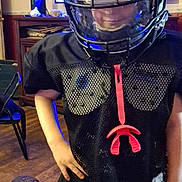 William is registered to the contest to win money with this photo: child, football_helmet, sports_gear, indoor, black_clothing, hand_on_hip, wooden_floor, television, furniture, hat, faceguard, silhouette_art, room, person, confident, standing, mesh_jersey, helmet_visor, casual, young