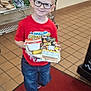 child, boy, glasses, red_shirt, spiderman, jeans, sneakers, tray, food, banana, cereal_box, cafeteria, floor_tiles, red_carpet, shelves, plastic_baskets, smiling, indoor, person, breakfast