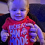adult_hand, baby, blue_light, casual, child, clothing, couch, cute, face, hand, happy, holding, indoors, infant, person, portrait, red_shirt, seated, smiling, text