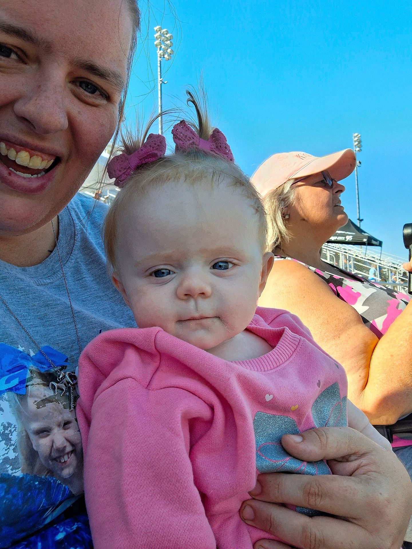 adult, baby, blue_sky, casual_clothing, child, daylight, family, glasses, hand, hat, outdoor, people, person, pink_bows, pink_sweater, portrait, smile, spectator, stadium, woman