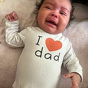 Aurora is registered to the contest to win money with this photo: baby, crying, infant, onesie, clothing, text, heart_symbol, blanket, soft_texture, person, child, indoor, expression, emotion, toddler, skin, hand, face, head, cute