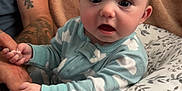 Weston is registered to the contest to win money with this photo: baby, child, pajamas, hand, tattoo, pillow, blanket, indoor, person, face, expression, holding, comfort, pattern, clothing, adult, skin, seated, closeup, home