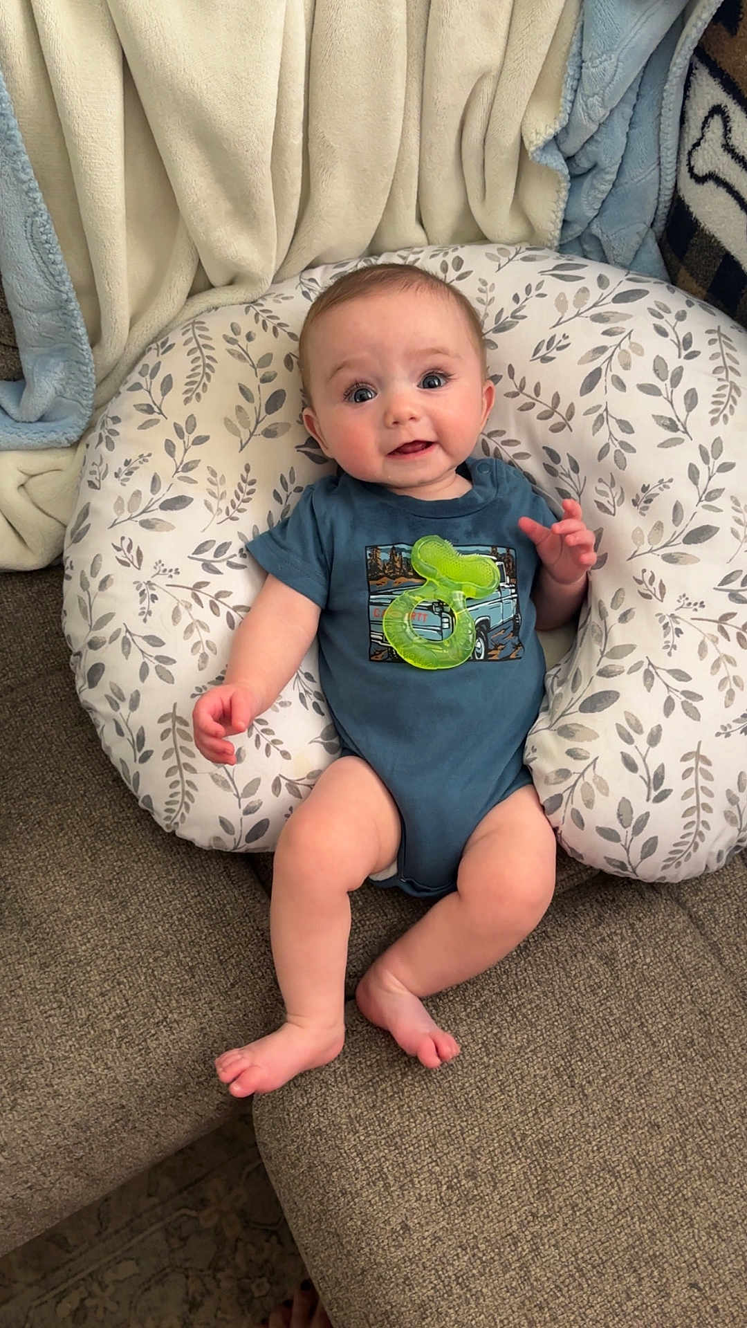 Weston is registered to the contest to win money with this photo: baby, infant, cushion, blanket, blue_onesie, teething_toy, couch, carpet, indoor, smiling, face, person, furniture, comfort, cute, child, relaxing, baby_gear, home, soft