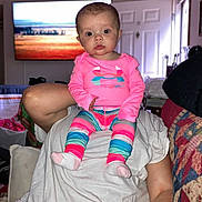 Gracie joined the competition — help win amazing prizes! baby, child, person, indoor, colorful_clothing, pink_shirt, striped_pants, socks, adult, shoulders, living_room, television, quilt, couch, curious, seated, portrait, cute, face, head