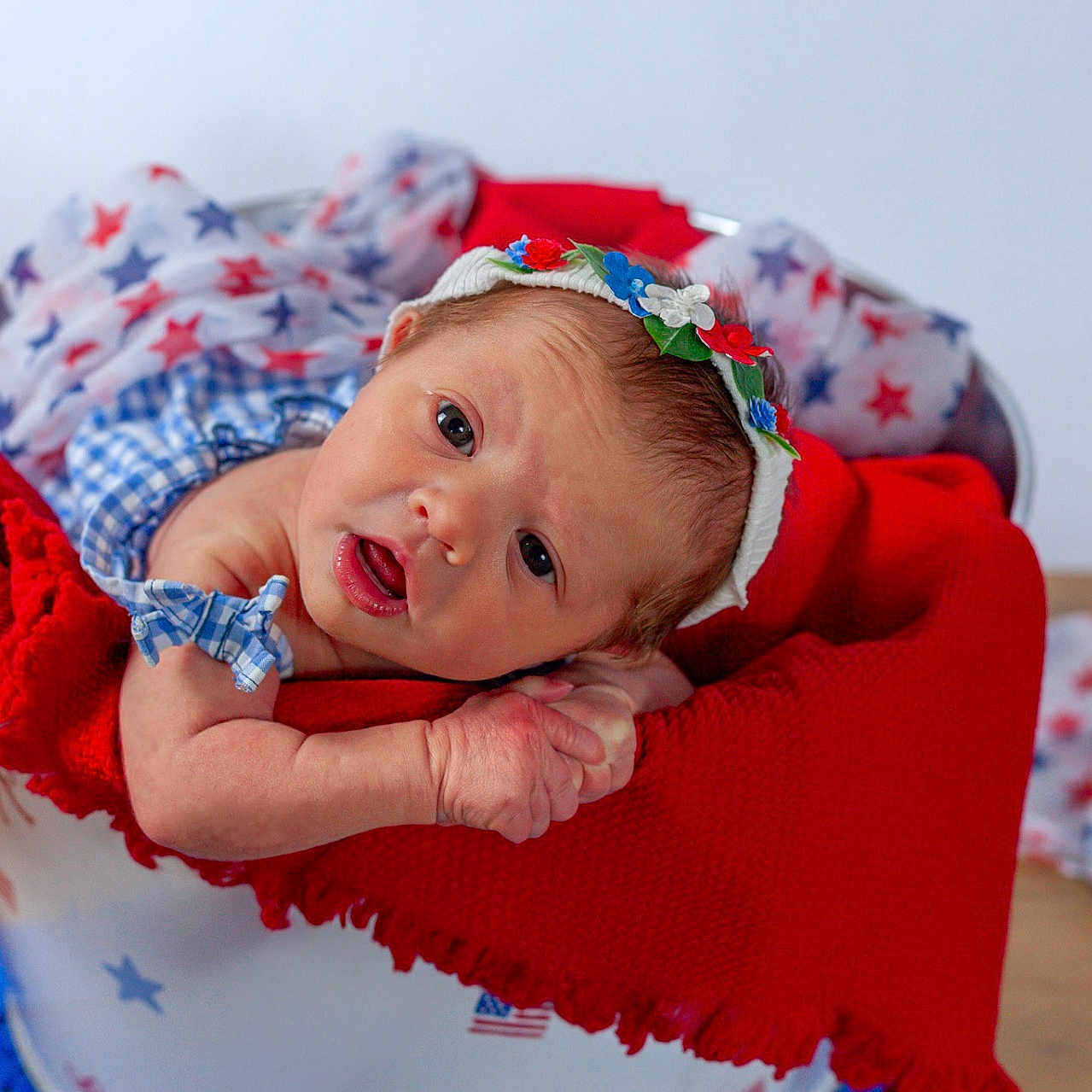 Harper joined the competition — help win amazing prizes! baby, newborn, infant, bucket, red_blanket, star_pattern, headband, blue_clothing, face, hands, wooden_surface, portrait, indoors, cute, expression, lying_down, colorful, decor, soft_texture, child