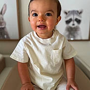 Jules a rejoint le concours — aidez-le/la à gagner de superbes lots ! toddler, child, smiling, indoor, portrait, white_tshirt, beige_pants, hands, face, eyes, hair, furniture, wall_art, animal_painting, raccoon, rabbit, cute, young_child, seated, cozy