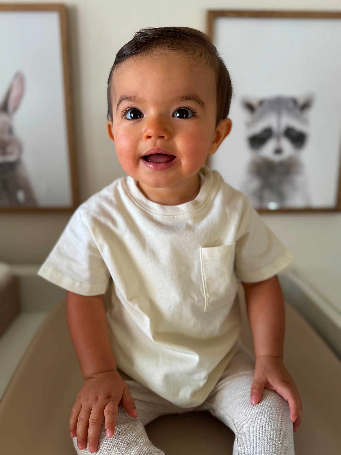 Jules a rejoint le concours — aidez-le/la à gagner de superbes lots ! toddler, child, smiling, indoor, portrait, white_tshirt, beige_pants, hands, face, eyes, hair, furniture, wall_art, animal_painting, raccoon, rabbit, cute, young_child, seated, cozy