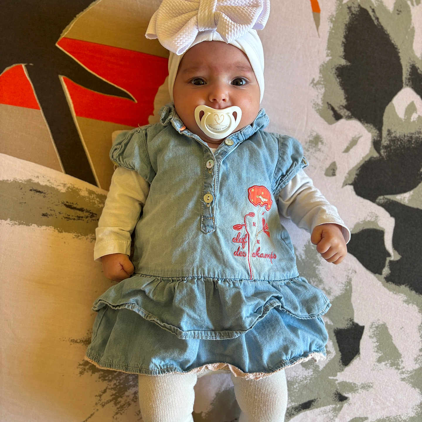 Inaya participe au concours pour gagner de l'argent avec cette photo : baby, baby_fashion, bow, child, clothing, colorful, cute, denim_dress, floral_pattern, headband, human, indoors, infant, lying_down, pacifier, patterned_background, pink_shoes, portrait, small_child, white_tights