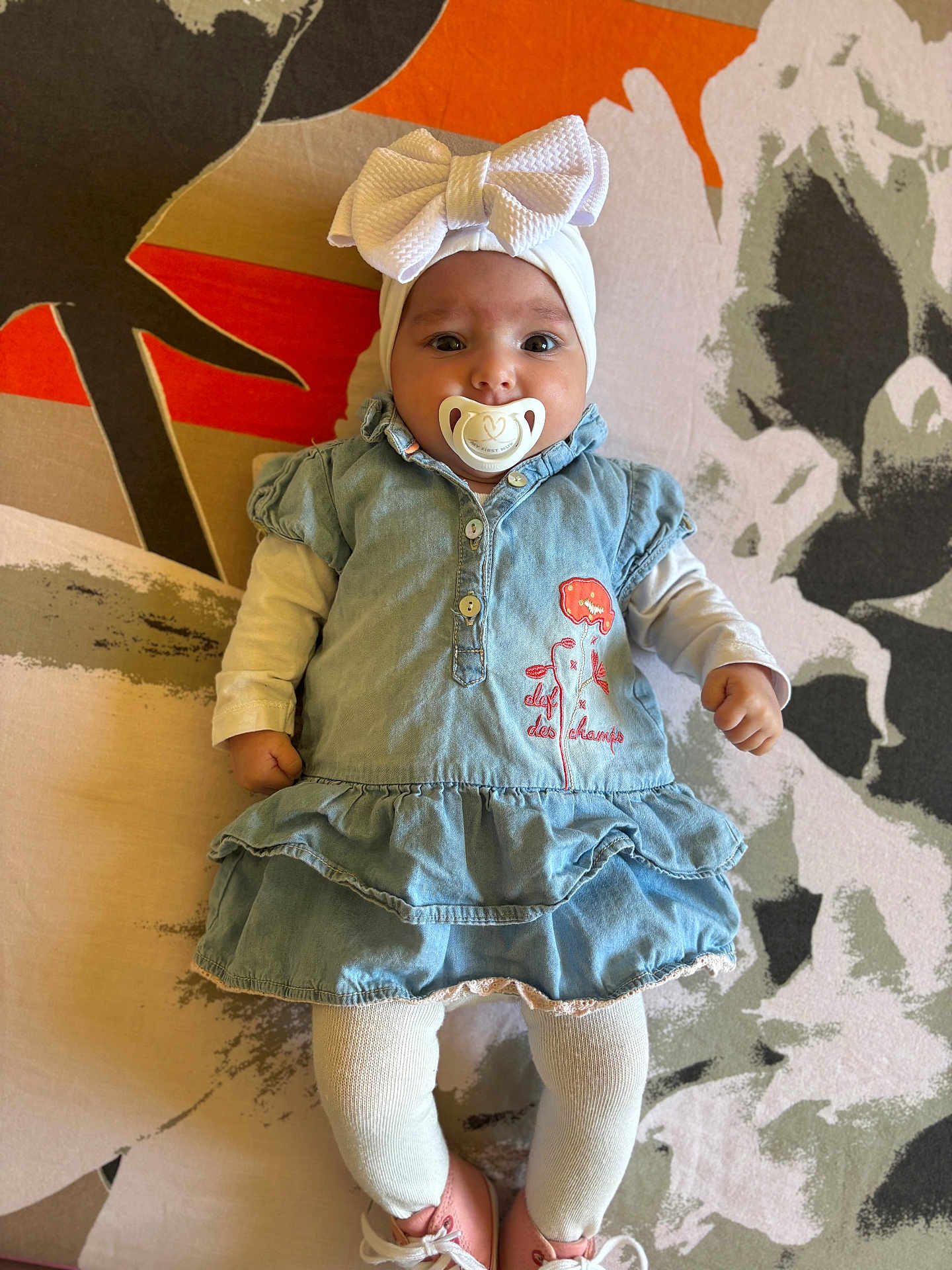 Inaya participe au concours pour gagner de l'argent avec cette photo : baby, pacifier, headband, bow, denim_dress, white_tights, pink_shoes, infant, child, cute, lying_down, patterned_background, colorful, floral_pattern, clothing, portrait, indoors, small_child, baby_fashion, human