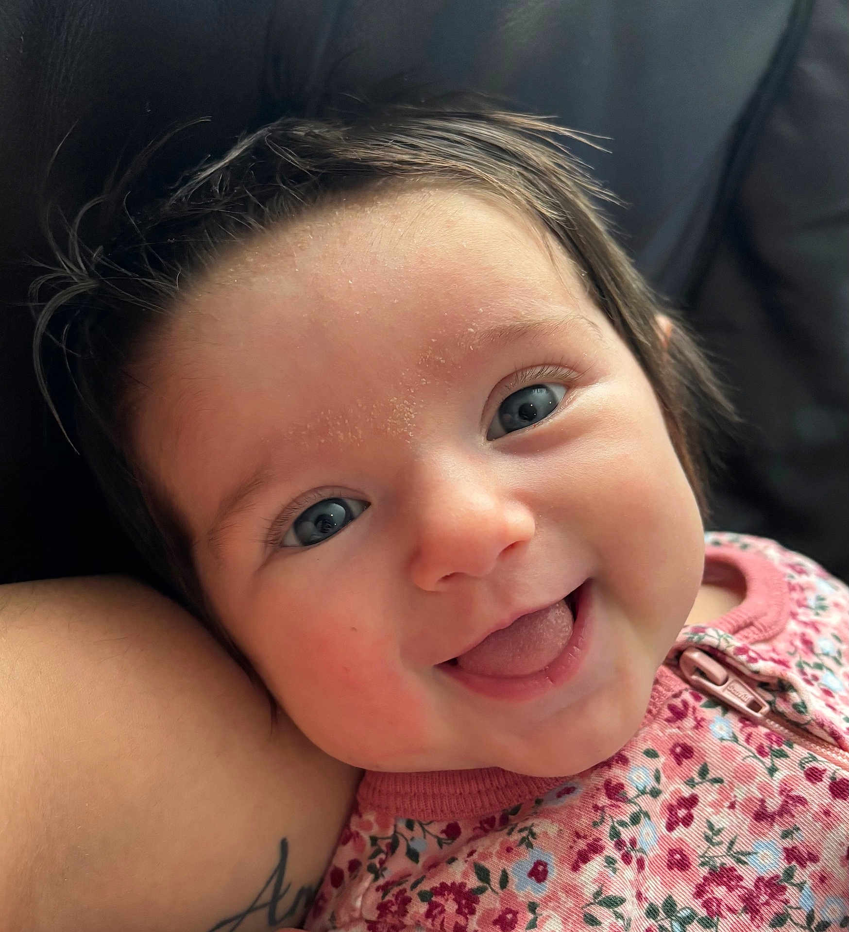 Eliana is registered to the contest to win money with this photo: baby, child, face, smile, tongue, blue_eyes, floral_clothing, infant, cute, closeup, skin, hair, cheeks, person, tattoo, arm, happy, expression, indoors, portrait
